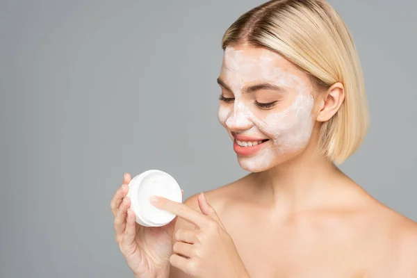 Femme blonde souriante avec masque facial tenant pot avec crème isolée sur gris — Photo de stock