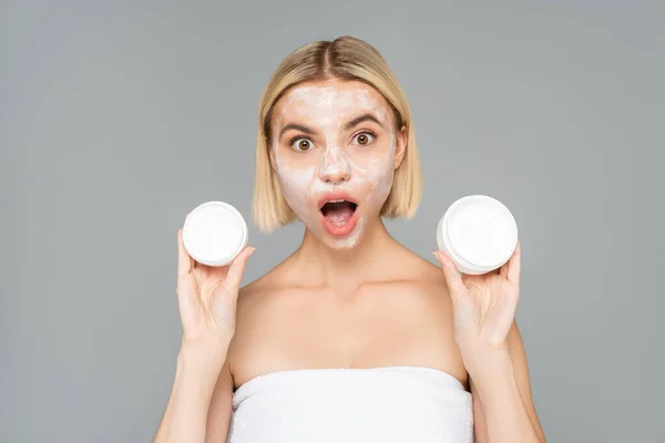 Amazed woman with facial mask holding jars with cosmetic cream isolated on grey — Stock Photo