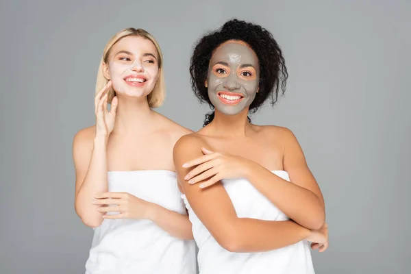 Smiling multicultural women in towels and facial masks isolated on grey — Stock Photo