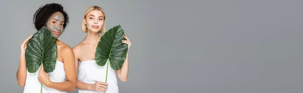 Multikulturelle Frauen in Handtüchern und Gesichtsmasken mit grünen Blättern auf grauem Banner — Stockfoto
