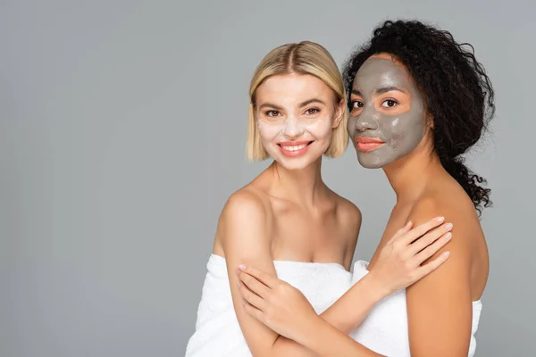 Mujeres sonrientes multiculturales con máscaras faciales abrazándose aisladas en gris - foto de stock