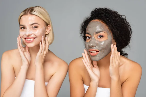 Cheerful multiethnic women in towels applying facial masks isolated on grey — Stock Photo