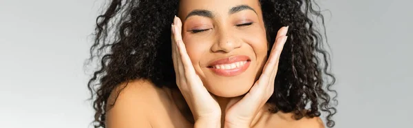 Donna afroamericana sorridente con le mani vicino al viso e gli occhi chiusi isolati su grigio, striscione — Foto stock