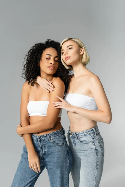 Blonde woman embracing african american woman in jeans on grey background — Stock Photo