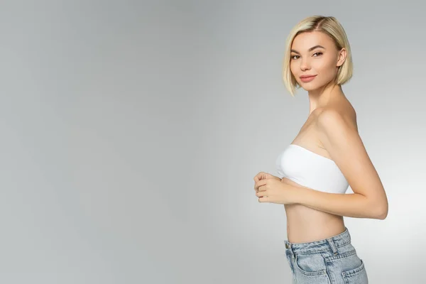 Mujer rubia en top blanco mirando a la cámara aislada en gris - foto de stock