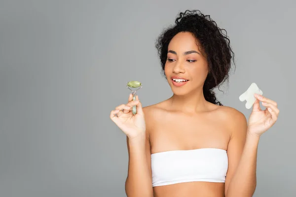 Smiling african american woman in white top holding jade roller and gua sha isolated on grey — Stock Photo