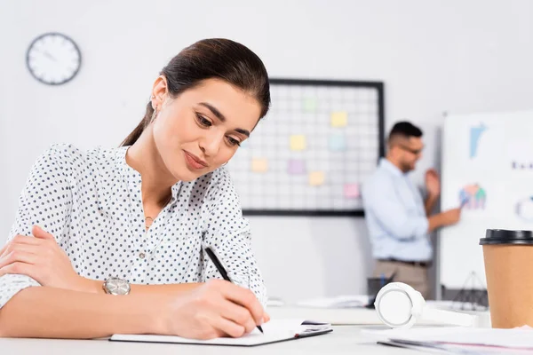 Cheerful businesswoman writing in notebook near paper cup on desk and coworker on blurred background — Stock Photo