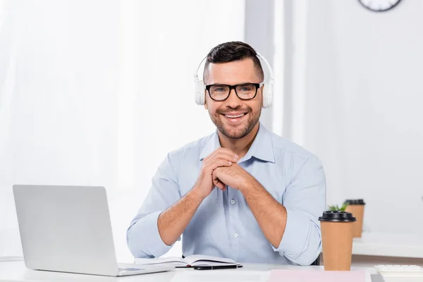 Hombre de negocios feliz en gafas y auriculares sonriendo mientras mira a la cámara - foto de stock