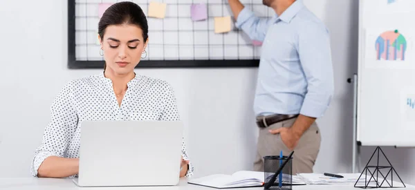 Mujer de negocios utilizando el ordenador portátil cerca de compañero de trabajo poner notas adhesivas a bordo sobre fondo borroso, bandera - foto de stock