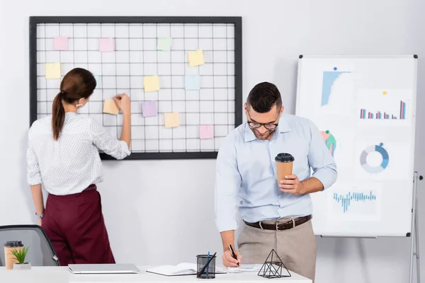 Businessman in glasses holding paper cup and writing in notebook near businesswoman putting sticky note on board — Stock Photo