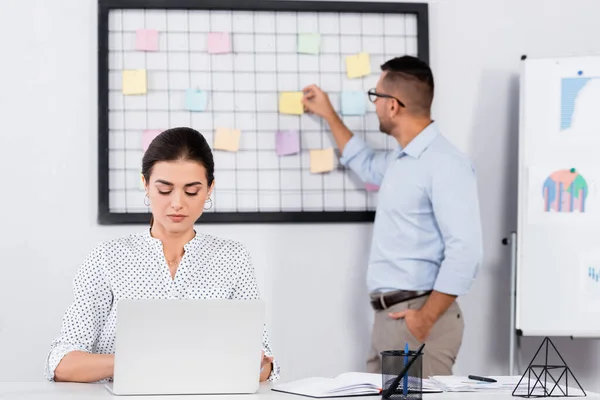 Businesswoman using laptop near coworker putting sticky notes on board and blurred background — Stock Photo