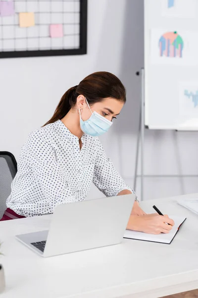 Бизнесвумен в медицинской маске пишет в блокноте рядом с ноутбуком — стоковое фото