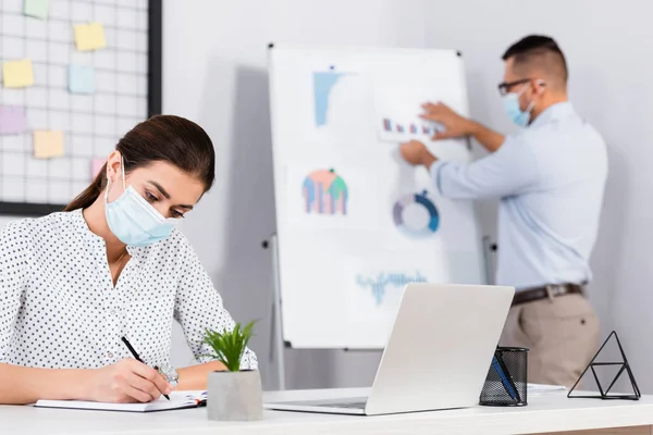 Businesswoman in medical mask writing in notebook near businessman on blurred background — Stock Photo