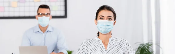 Mujer de negocios en máscara médica mirando a la cámara cerca de hombre de negocios sobre fondo borroso, pancarta - foto de stock