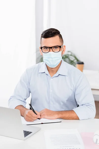 Geschäftsmann mit medizinischer Maske und Brille blickt in die Kamera und hält Stift in der Nähe von Notizbuch und Laptop auf dem Schreibtisch — Stockfoto