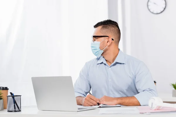 Hombre de negocios en máscara médica y gafas mirando hacia otro lado mientras sostiene la pluma cerca de portátil y portátil en el escritorio - foto de stock