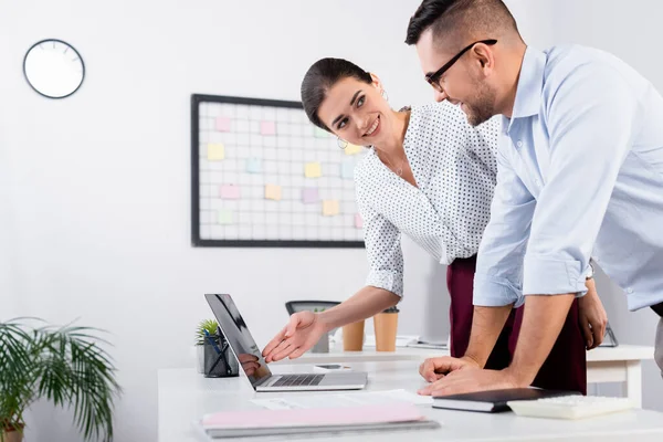 Mujer de negocios feliz apuntando con la mano a la computadora portátil con pantalla en blanco en el escritorio - foto de stock