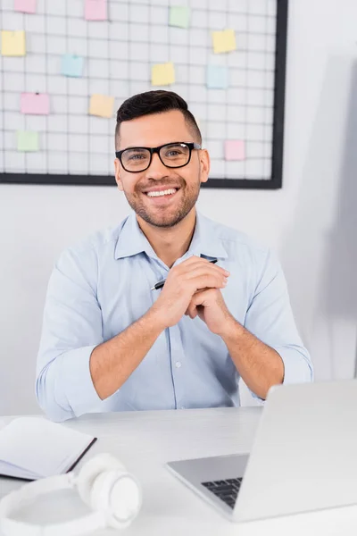 Alegre hombre de negocios en gafas sonriendo cerca de la computadora portátil en el escritorio - foto de stock