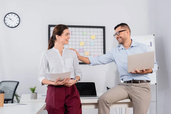 Happy businessman in glasses holding laptop and touching shoulder of coworker with documents — Stock Photo