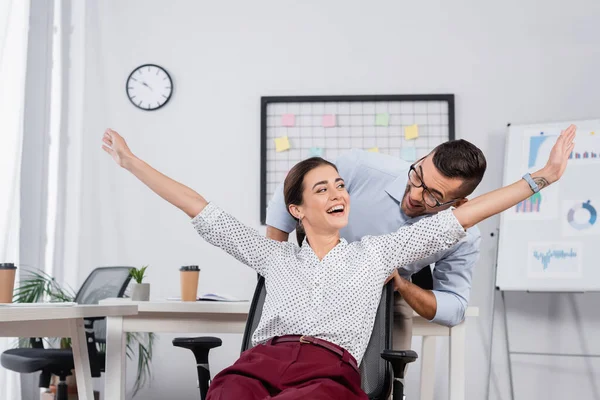 Businessman behind excited businesswoman with outstretched hands on chair — Stock Photo