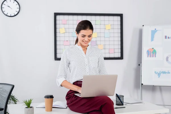 Femme d'affaires gaie regardant ordinateur portable et assis sur le bureau — Photo de stock