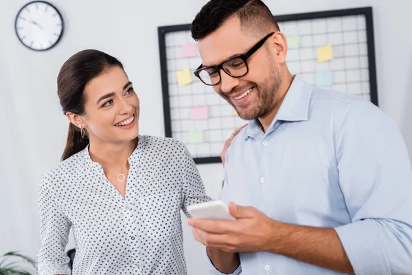 Mujer de negocios feliz mirando a hombre de negocios con teléfono inteligente en la oficina - foto de stock