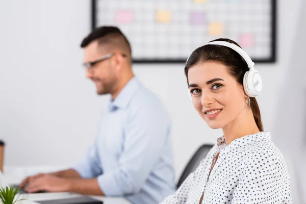 Feliz mujer de negocios en auriculares inalámbricos mirando a la cámara cerca de hombre de negocios en un fondo borroso - foto de stock