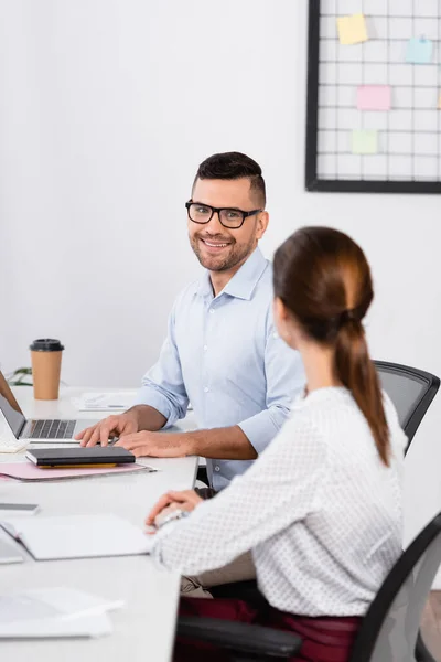 Empresário alegre em óculos olhando para empresária sentada na mesa em primeiro plano borrado — Fotografia de Stock