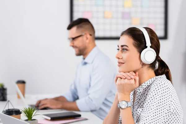 Pensativa mujer de negocios en auriculares inalámbricos cerca de hombre de negocios que trabaja en primer plano borrosa - foto de stock
