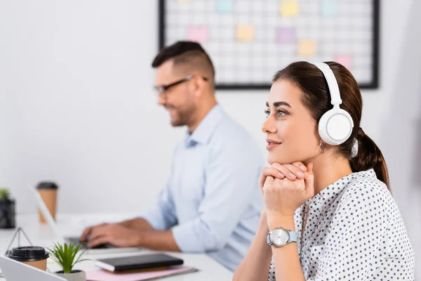 Nachdenkliche Geschäftsfrau mit drahtlosen Kopfhörern in der Nähe von Geschäftsleuten, die an verschwommenem Vordergrund arbeiten — Stockfoto