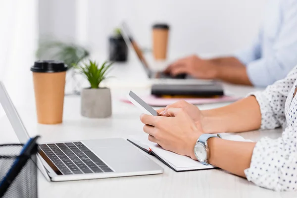 Cropped view of businessman holding smartphone near laptop on desk — Stock Photo