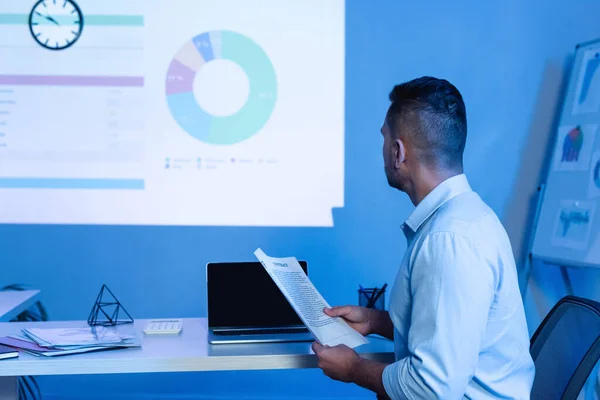 Empresario celebración de contrato cerca de la computadora portátil con pantalla en blanco mientras mira gráficos y gráficos en la pared - foto de stock