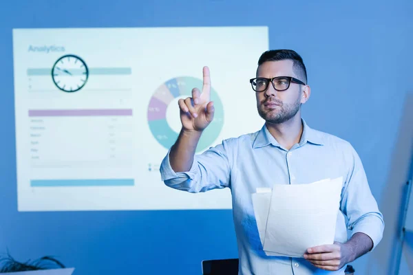 Geschäftsmann in Brille zeigt mit dem Finger und hält Papiere in der Nähe von Diagrammen an der Wand — Stockfoto