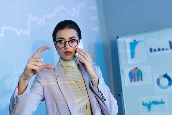 Бізнес-леді в окулярах жестикулює біля цифрових графіків і розмовляє на смартфоні — стокове фото