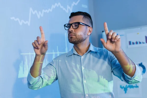 Hombre de negocios en gafas apuntando con los dedos cerca de gráficos digitales en la oficina - foto de stock