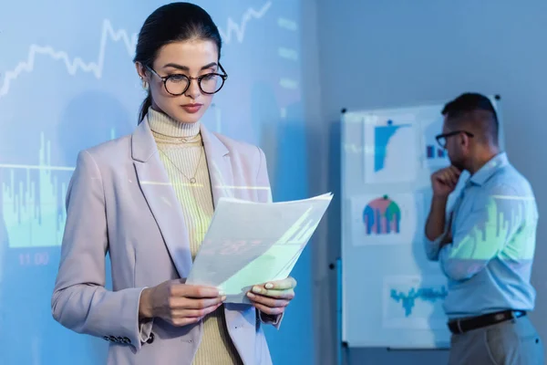 Mujer de negocios en gafas mirando documentos cerca de compañero de trabajo en fondo borroso - foto de stock