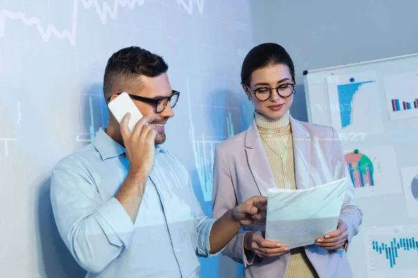 Alegre hombre de negocios hablando en el teléfono inteligente y mirando los documentos cerca de compañero de trabajo en gafas - foto de stock