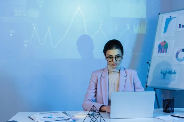 Geschäftsfrau in Brille mit Laptop nahe digitaler Graphik an Wand — Stockfoto