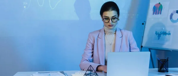 Mujer de negocios en gafas usando el ordenador portátil cerca de gráficos digitales en la pared, bandera - foto de stock