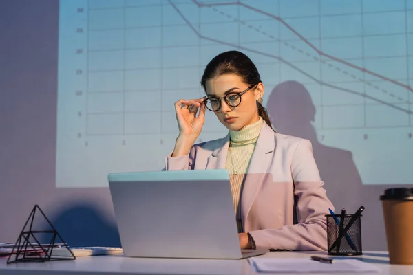 Businesswoman adjusting glasses and using laptop near digital graphs on wall and paper cup on blurred foreground — Stock Photo