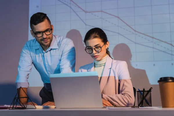 Business people in glasses looking at laptop with digital charts on wall on background — Stock Photo