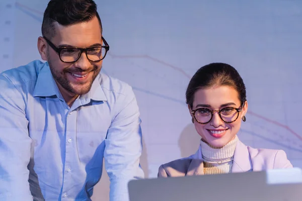 Cheerful business people in glasses looking at laptop near digital charts on wall — Stock Photo