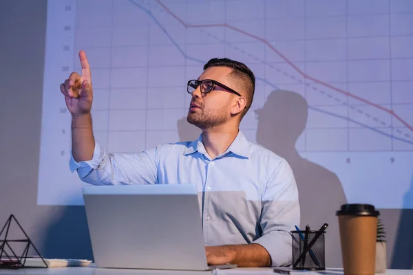 Businessman in glasses pointing with finger near laptop and graphs on wall — Stock Photo