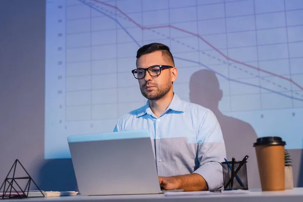 Homme d'affaires en lunettes regardant ordinateur portable près des graphiques sur le mur et tasse en papier sur le bureau — Photo de stock