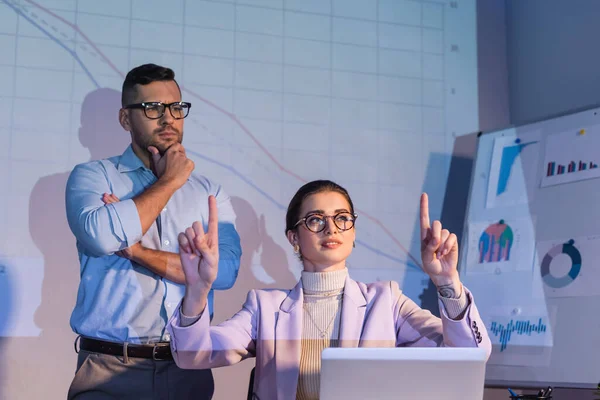 Empresaria en gafas apuntando con los dedos cerca de hombre de negocios en gafas mirando gráficos digitales — Stock Photo