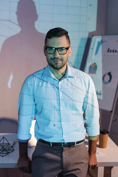 Uomo d'affari sorridente in occhiali in piedi vicino alla scrivania con tazza di carta — Foto stock