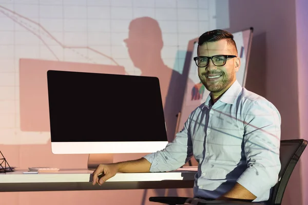 Felice uomo d'affari in occhiali guardando la fotocamera mentre seduto vicino al monitor del computer con schermo vuoto — Foto stock