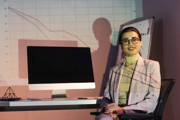 Femme d'affaires heureuse dans des lunettes regardant la caméra tout en étant assis près de l'écran de l'ordinateur avec écran blanc — Photo de stock