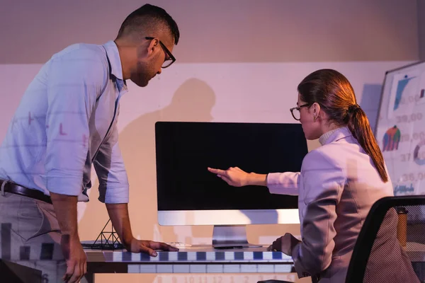 Businesswoman in glasses pointing with finger at computer monitor with blank screen near coworker in office — Stock Photo