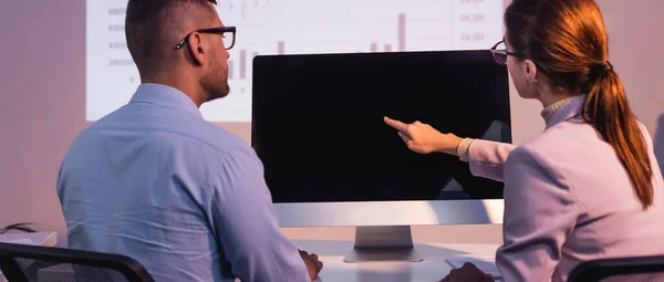 Empresária em óculos apontando com o dedo para monitor de computador com tela em branco perto colega de trabalho no escritório, banner — Fotografia de Stock